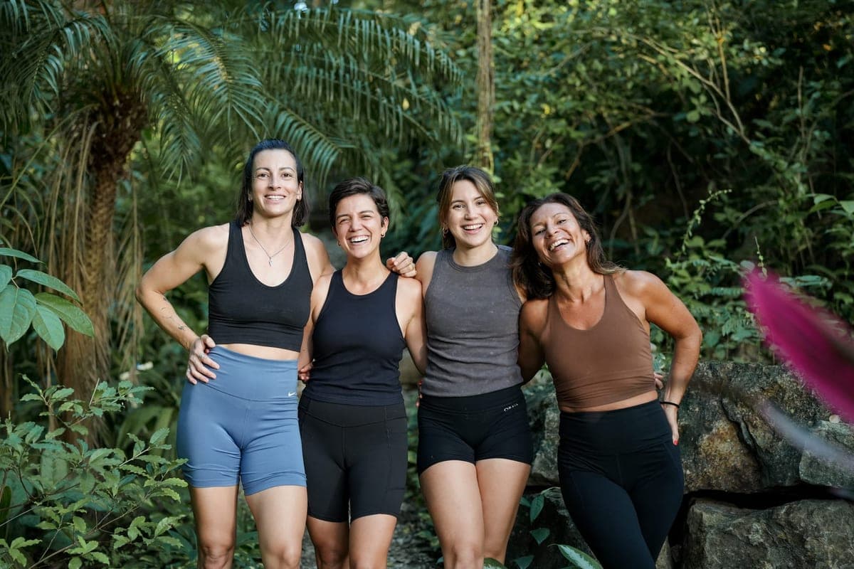 Four women stand together in the jungle with their arms around each other, smiling.
