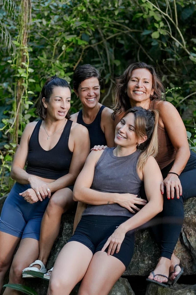 Four women sit close together on a rock in the jungle, laughing.