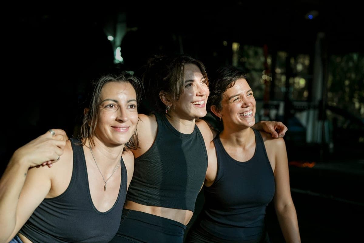 Three women with their arms around each other smile in the dim studio.