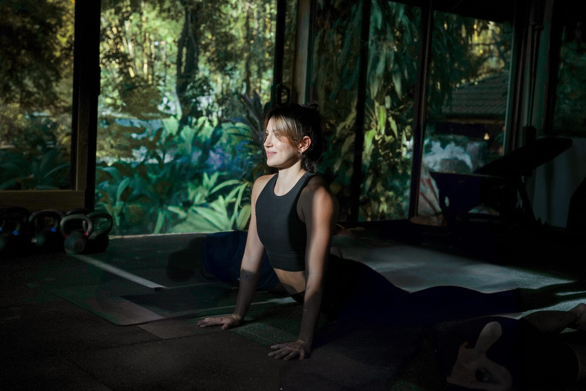Inside the studio, a woman smiles as she lifts into a stretch on her mat, with the jungle filling the glass walls behind her.